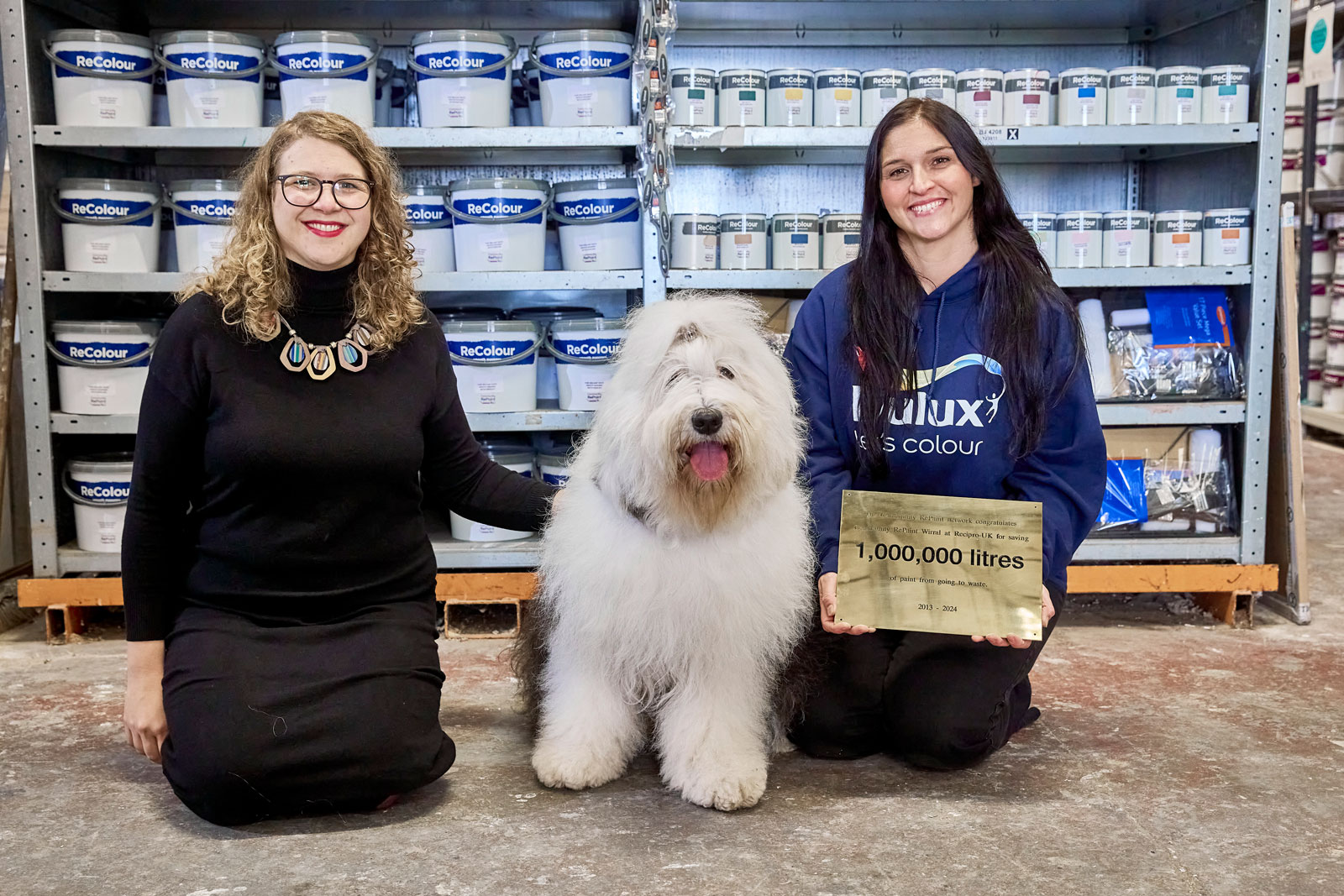 Two people with an old English sheepdog in the middle of them. Some tins of paint on shelves behind them