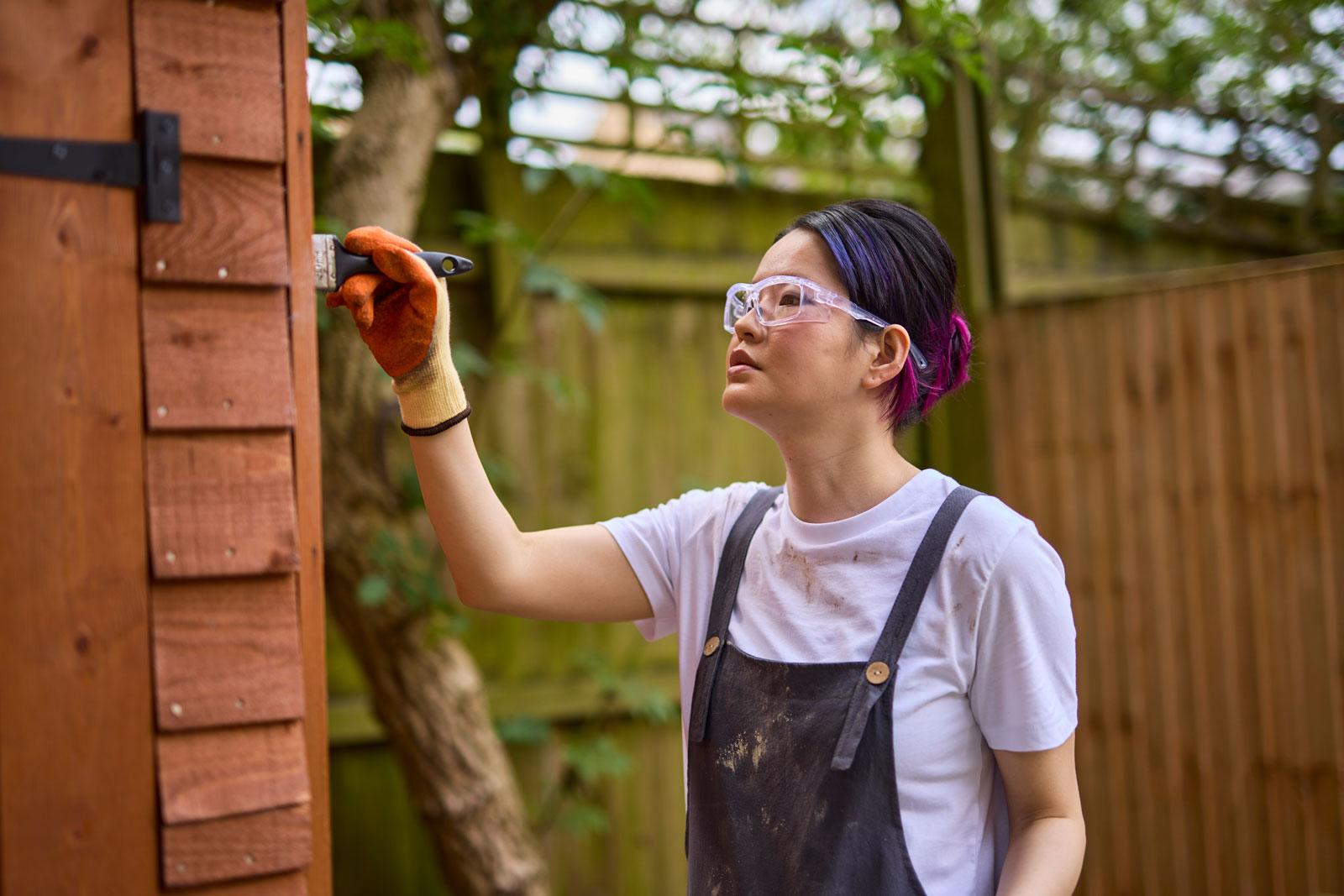 Photograph of a woman painting a shed using safety glasses